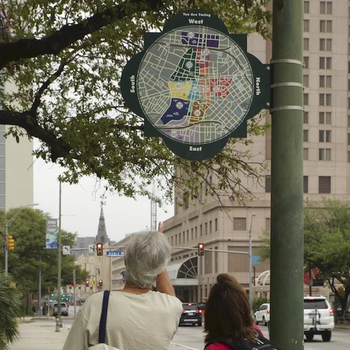 Two people looking up at a circular wayfinding map of San Antonio affixed on a lamp-post. Shows cardinal direction and neighbourhoods in different colours